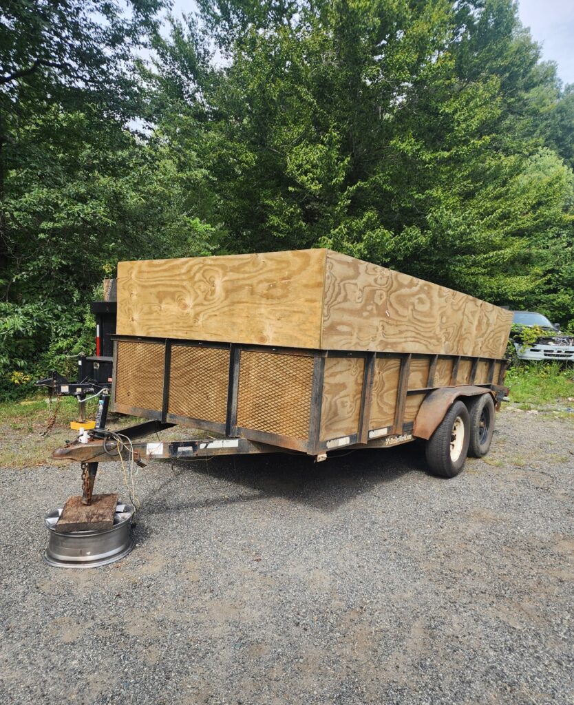 Utility trailer with wooden side panels is parked on gravel near trees, with a wheel rim under its hitch for support.