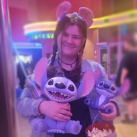 Woman smiling at a theater, holding popcorn and two Stitch plush toys, wearing Stitch ears headband.