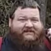 A bearded man with short hair stands outdoors in front of greenery, looking at the camera.