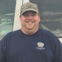 Man wearing a camouflage cap and navy shirt standing in front of a vehicle, smiling at the camera.