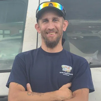 Man with a beard, wearing a navy shirt and hat with sunglasses, stands smiling in front of a white vehicle.