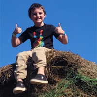 Child sitting on a haystack giving two thumbs up, wearing a black shirt and tan pants, against a clear blue sky.