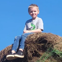 Young boy sits on a hay bale, wearing jeans and a grey t-shirt, with a clear blue sky in the background.
